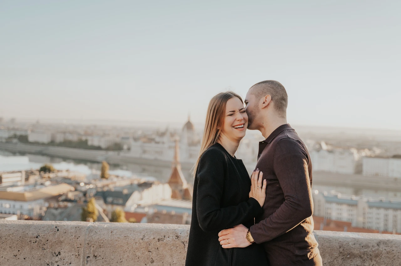 Laughter with a Budapest skyline backdrop at a Budapest couple photoshoot A landscape portrait of the man kissing his laughing partner near her temple with Budapest’s rooftops and Parliament behind them during their couple photoshoot in Budapest.