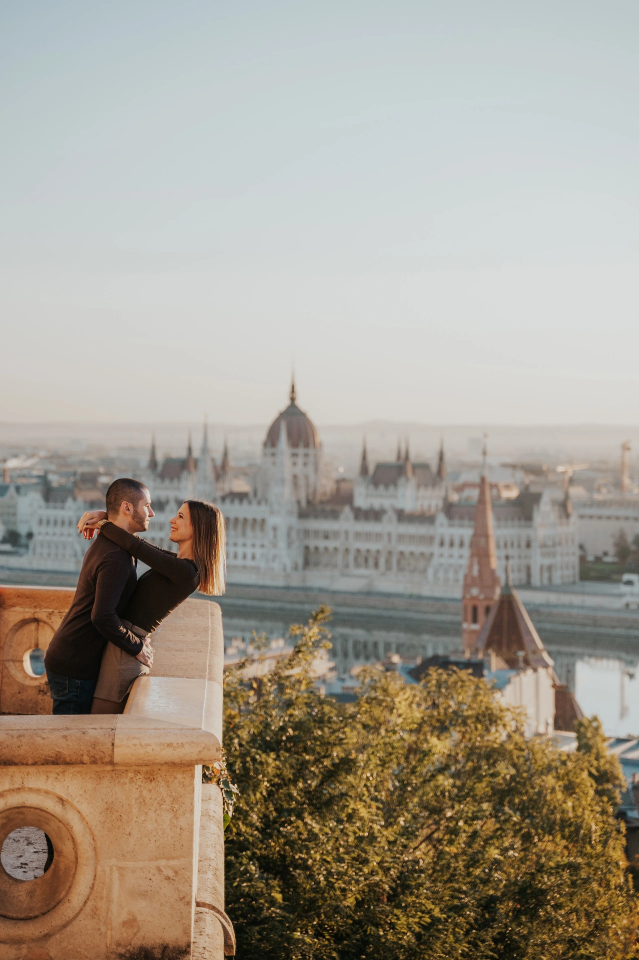 The couple embracing beside a carved balcony at Fisherman’s Bastion with Parliament across the river behind them during their couple photoshoot in Budapest.