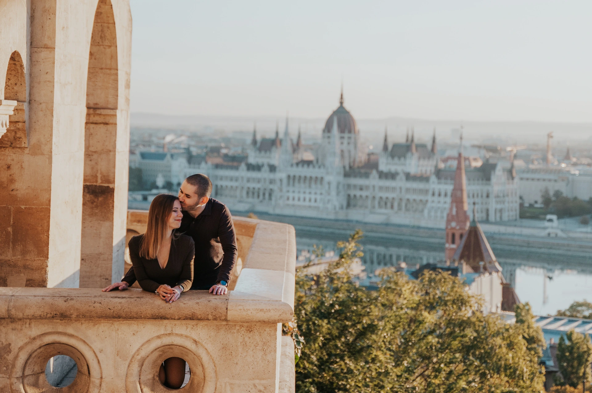 A kiss above the Danube and Parliament at a Budapest couple photoshoot A landscape portrait of the man kissing his partner near her temple at Fisherman’s Bastion’s balcony with Parliament across the river behind them during their couple photoshoot in Budapest.