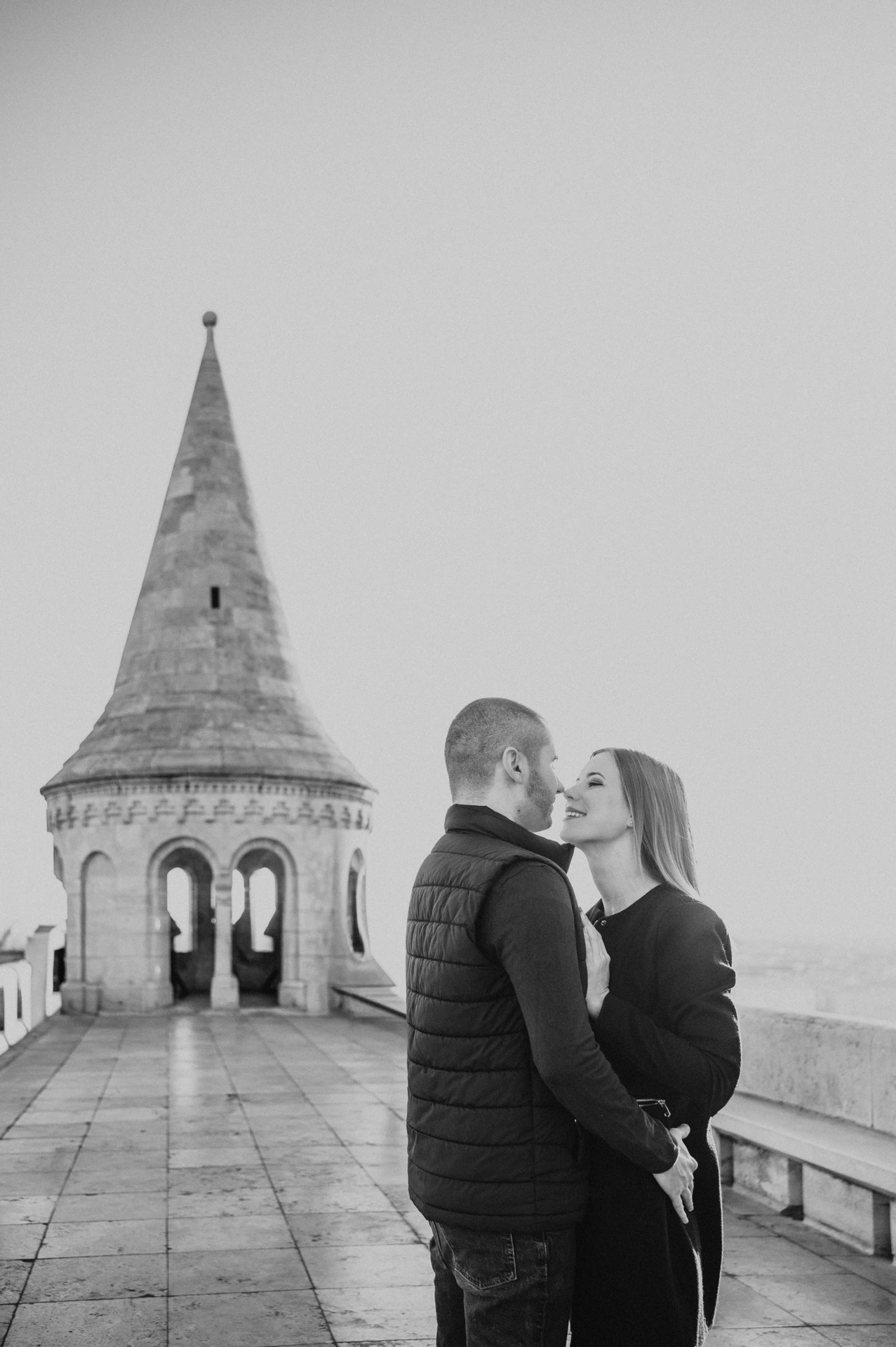 Black-and-white portrait of the couple embracing beside Fisherman’s Bastion’s pointed stone tower during their couple photoshoot in Budapest.