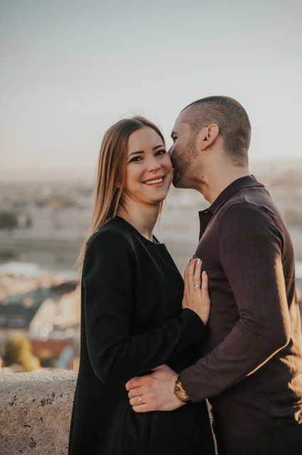 A little laughter above the city at a Budapest couple photoshoot A close portrait of the man kissing his smiling partner near her temple on a terrace overlooking Budapest during their couple photoshoot in Budapest.