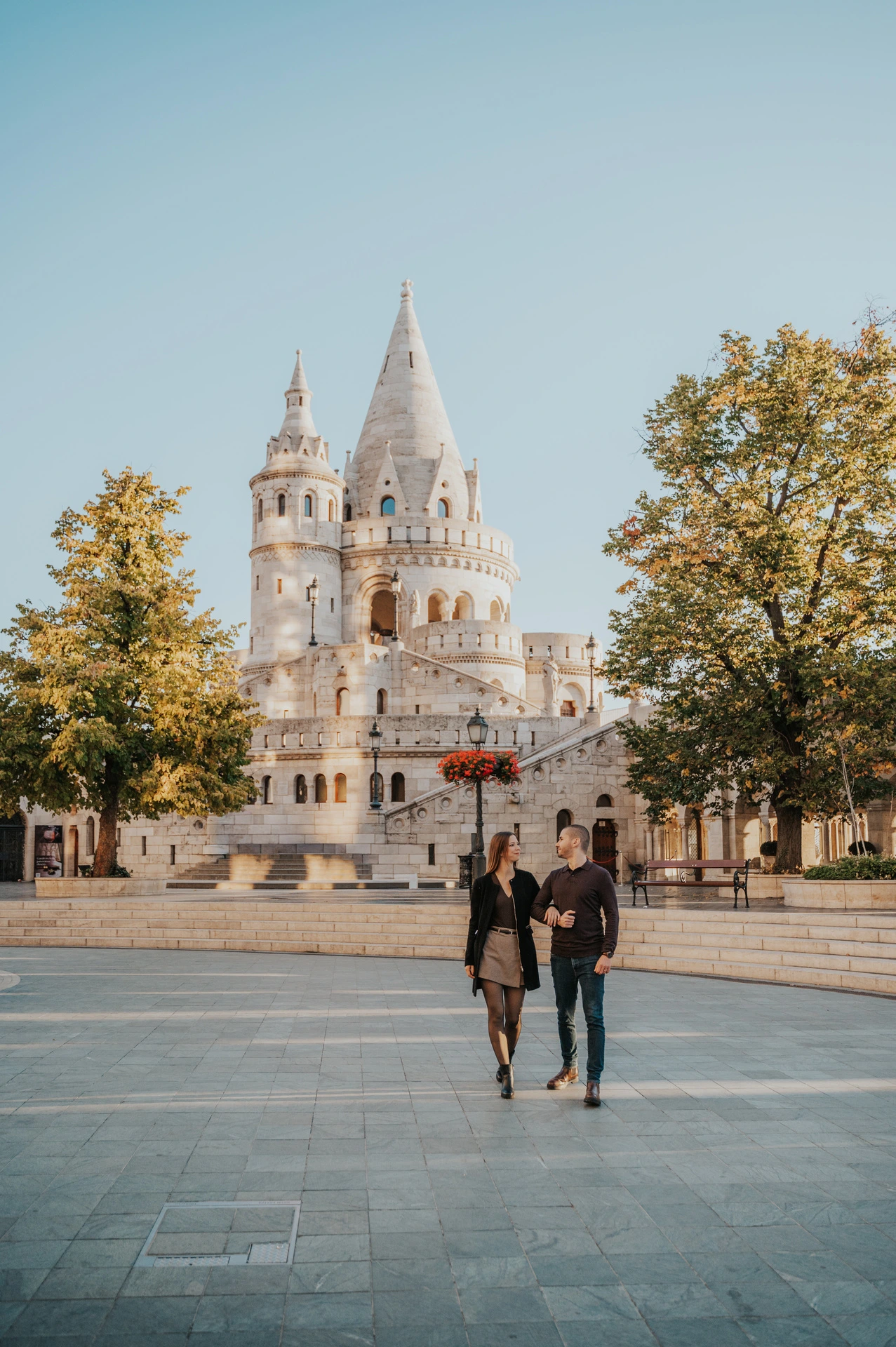A wide portrait of the couple walking arm in arm across the square with Fisherman’s Bastion’s towers and autumn trees behind them during their couple photoshoot in Budapest.