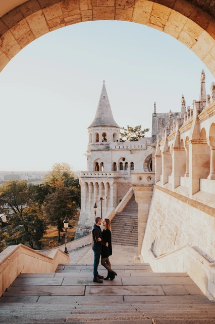 A romantic portrait on the stone stairs at a Budapest couple photoshoot The couple embracing on the broad stone stairs beneath an arch at Fisherman’s Bastion with a pointed tower behind them during their couple photoshoot in Budapest.