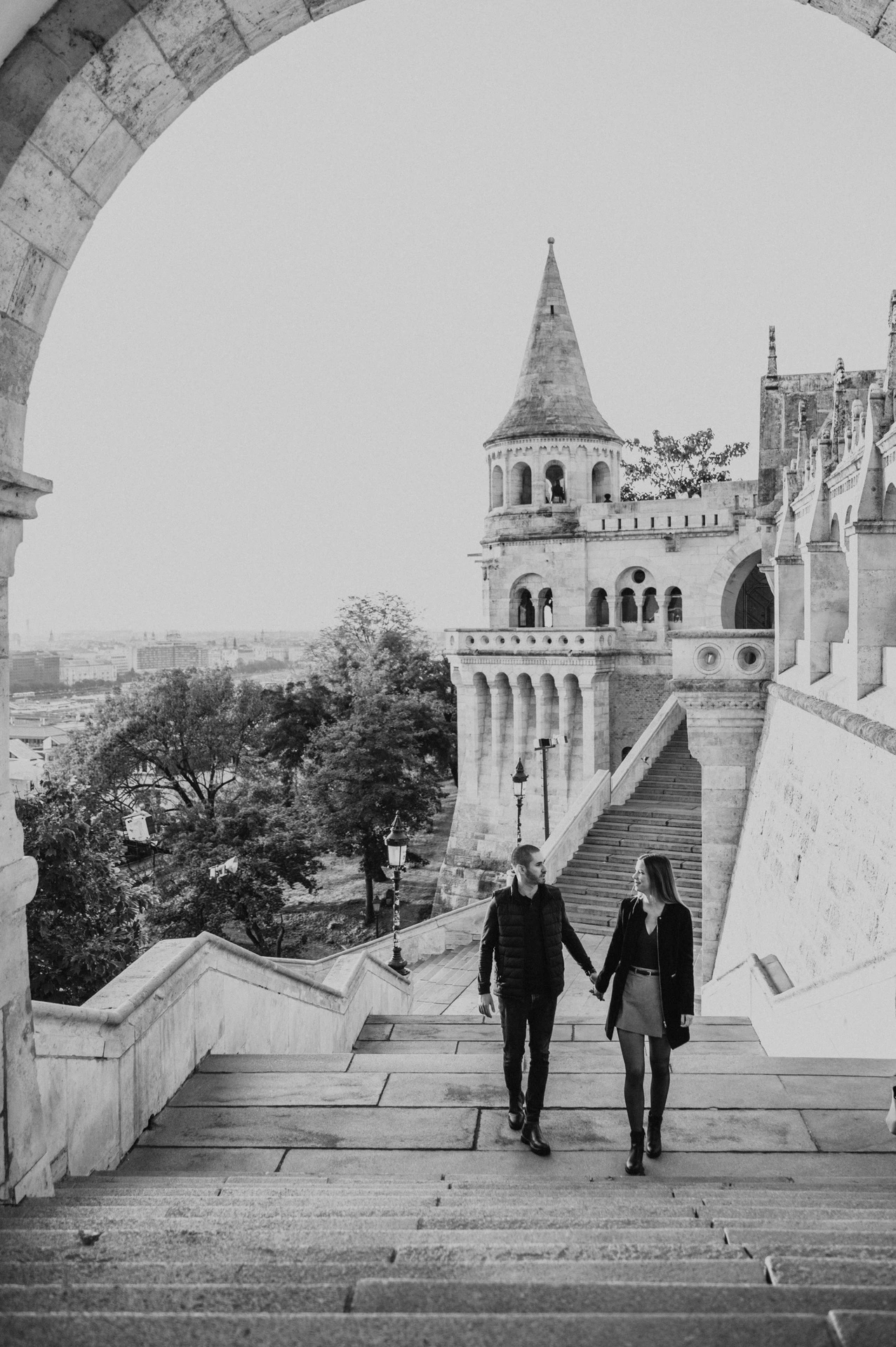 Black-and-white portrait of the couple walking up Fisherman’s Bastion’s broad stone steps with its tower behind them during their couple photoshoot in Budapest.
