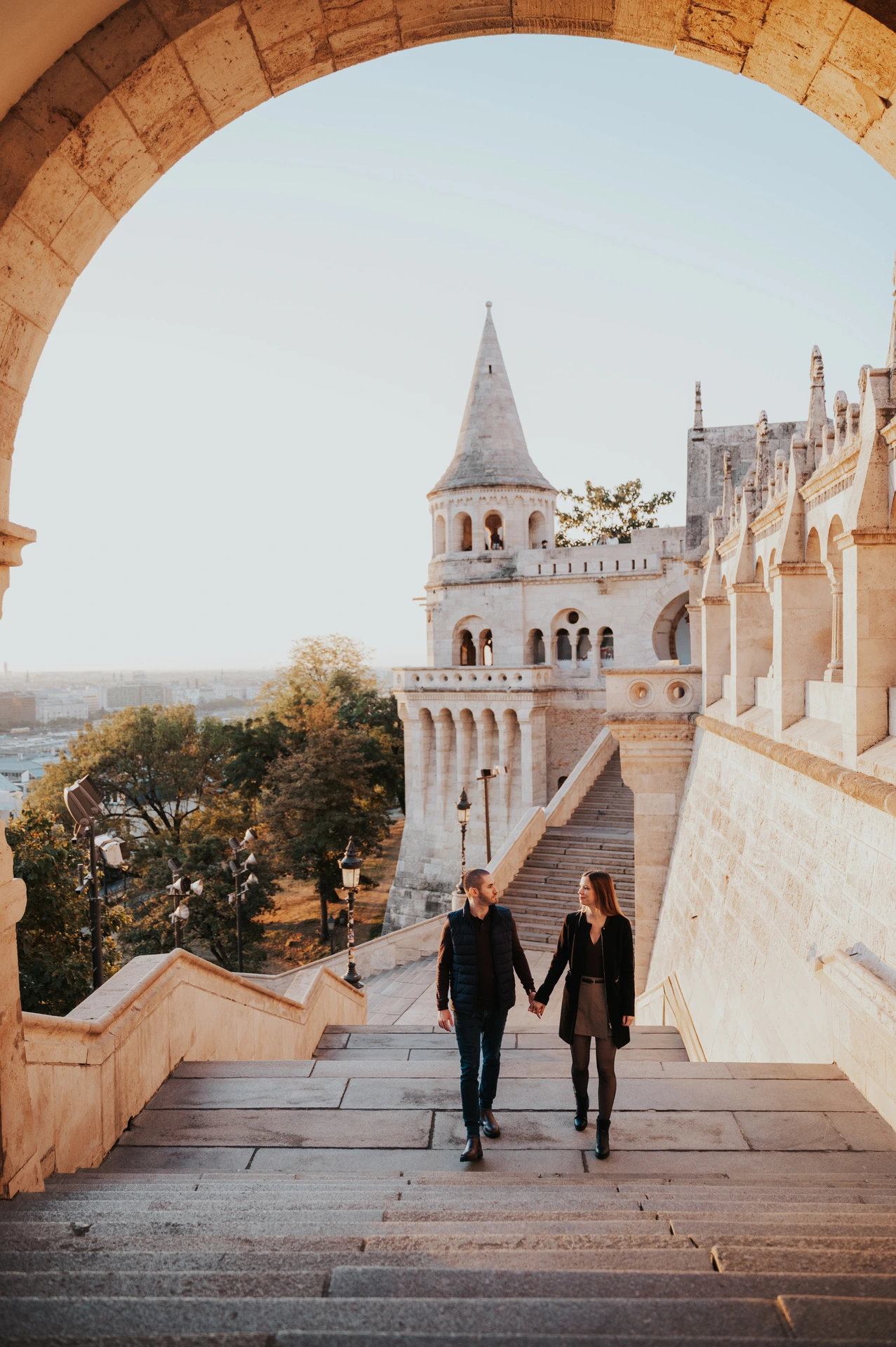 The couple walking hand in hand up the broad stone steps at Fisherman’s Bastion beneath a large arch during their couple photoshoot in Budapest.