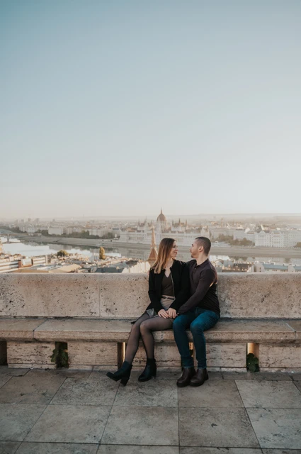 Time together on a bench above the city at a Budapest couple photoshoot The couple sitting together on a stone bench with Parliament and Budapest’s rooftops behind them at Fisherman’s Bastion during their couple photoshoot in Budapest.