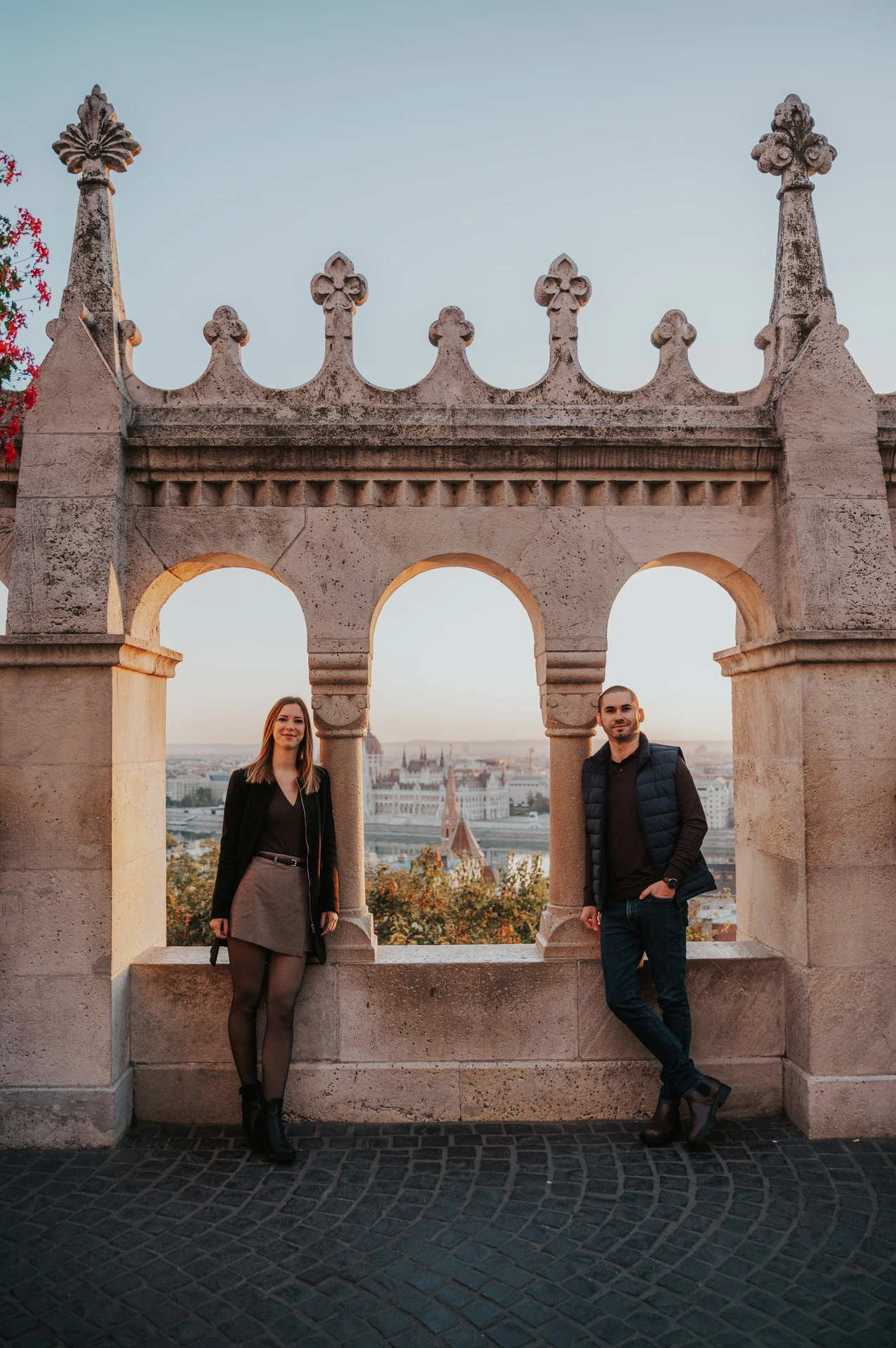The couple standing apart beneath three stone arches at Fisherman’s Bastion with Parliament across the Danube behind them during their couple photoshoot in Budapest.