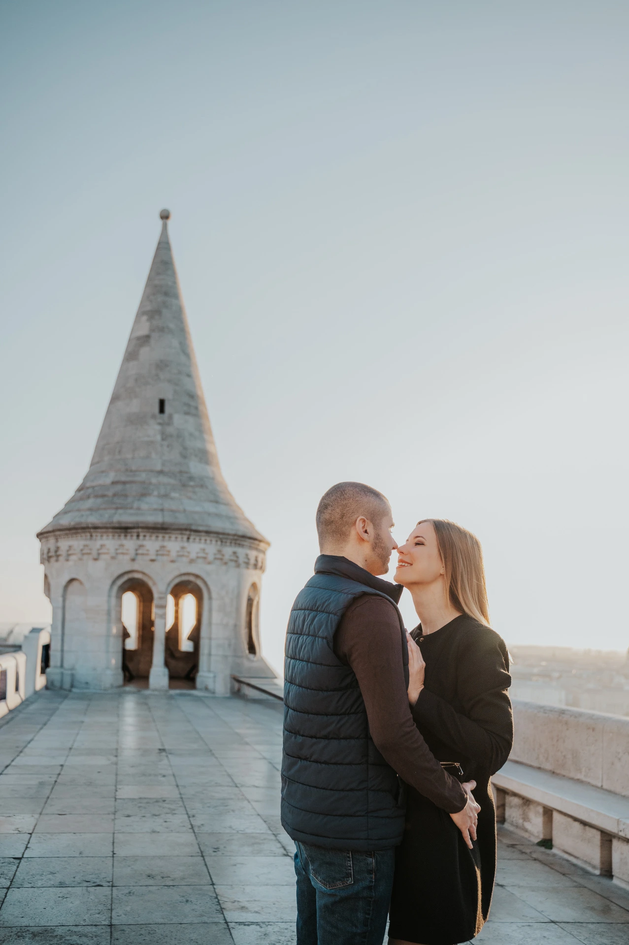 The couple embracing on the stone terrace with a pointed tower at Fisherman’s Bastion behind them during their couple photoshoot in Budapest.