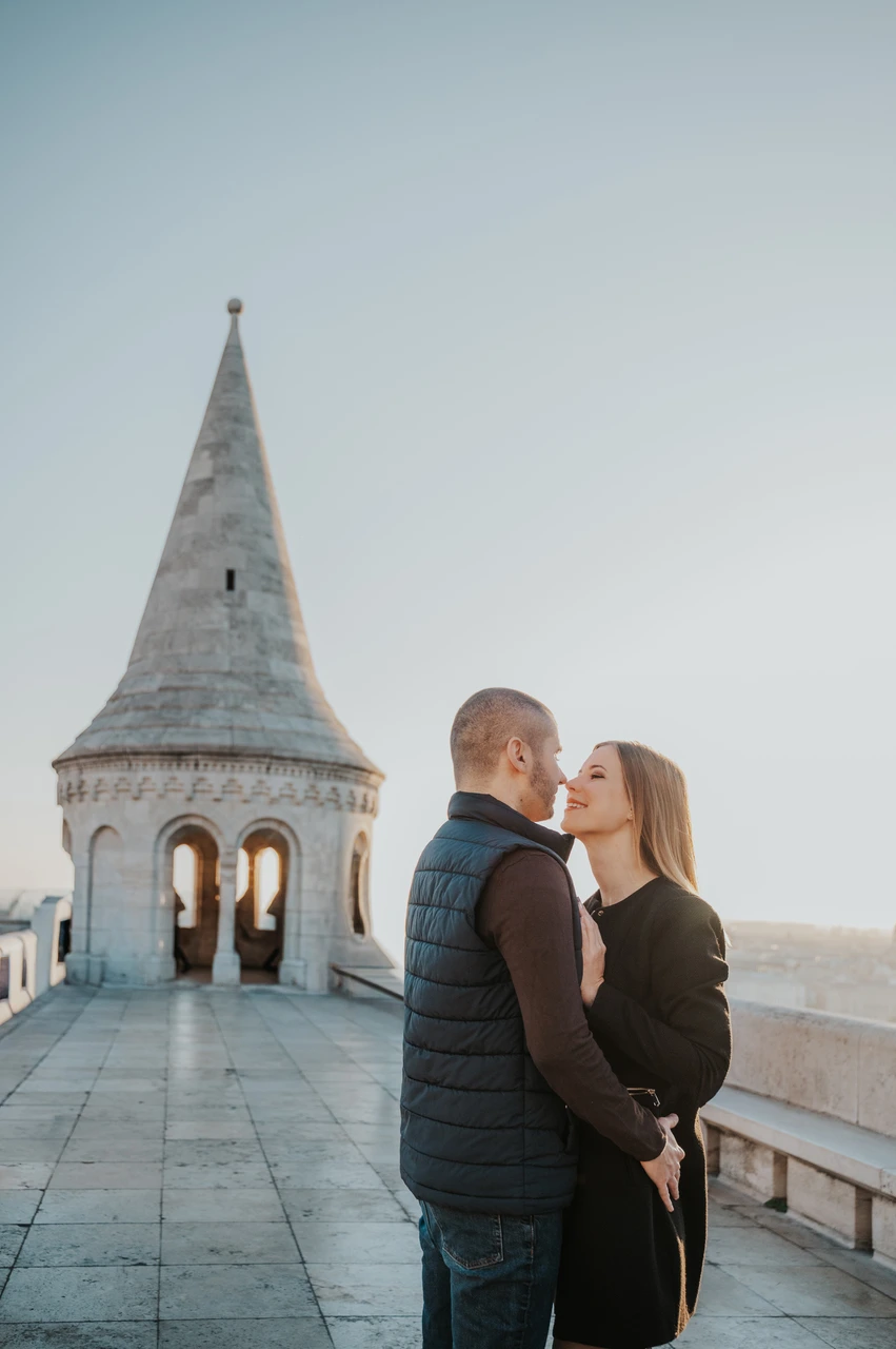 A romantic portrait beside the Bastion tower at a Budapest couple photoshoot The couple embracing on the stone terrace with a pointed tower at Fisherman’s Bastion behind them during their couple photoshoot in Budapest.
