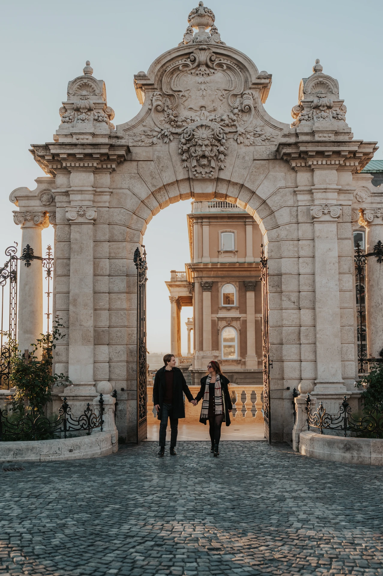 Couple holding hands beneath the ornate Buda Castle gate during their morning photoshoot in Budapest