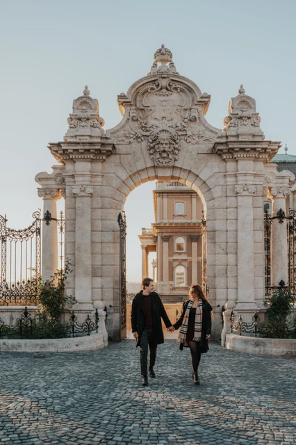 Budapest couple photoshoot walking through the castle gate Couple walking towards the camera through the ornate Buda Castle gate during their Budapest photoshoot