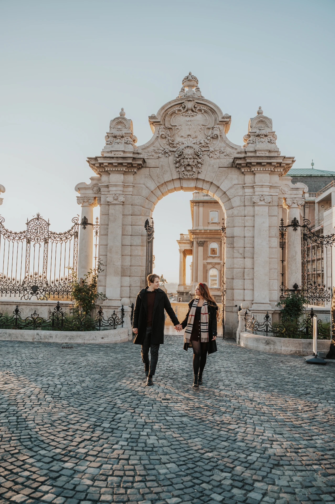 Couple walking across the cobbles beneath the decorated Buda Castle gate during their morning Budapest photoshoot
