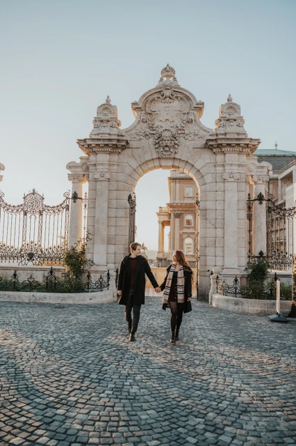 Morning couple photoshoot in Budapest around the castle gate Couple walking across the cobbles beneath the decorated Buda Castle gate during their morning Budapest photoshoot