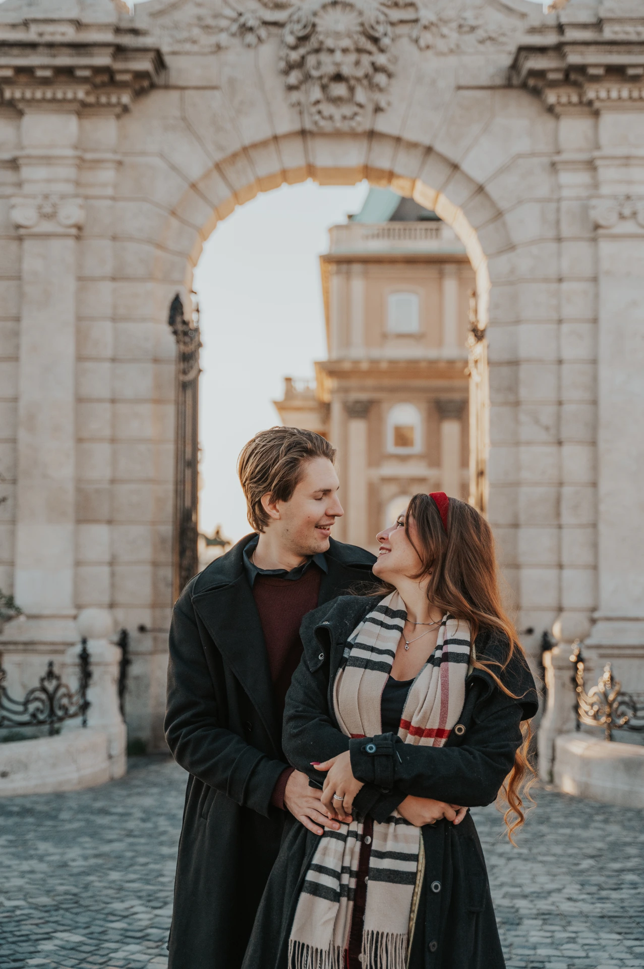 Couple smiling at each other while embracing beneath the Buda Castle arch during their photoshoot in Budapest