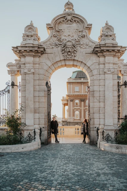 Budapest couple photoshoot beneath the stone castle arch Couple standing close together beneath the stone arch of the Buda Castle gate during their photoshoot in Budapest