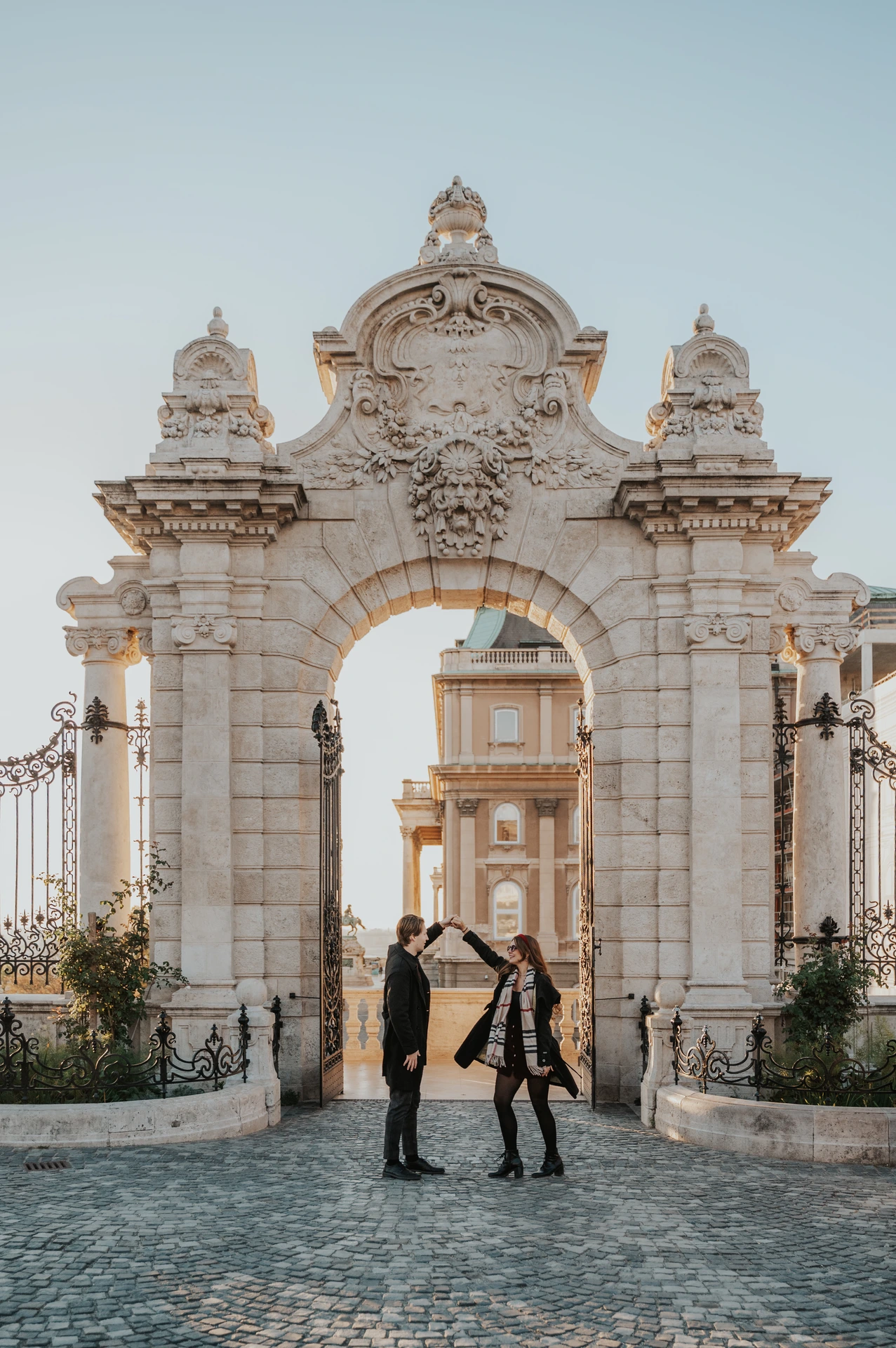 Man lifting their joined hands as his partner turns beside the Buda Castle gate during their couple photoshoot in Budapest