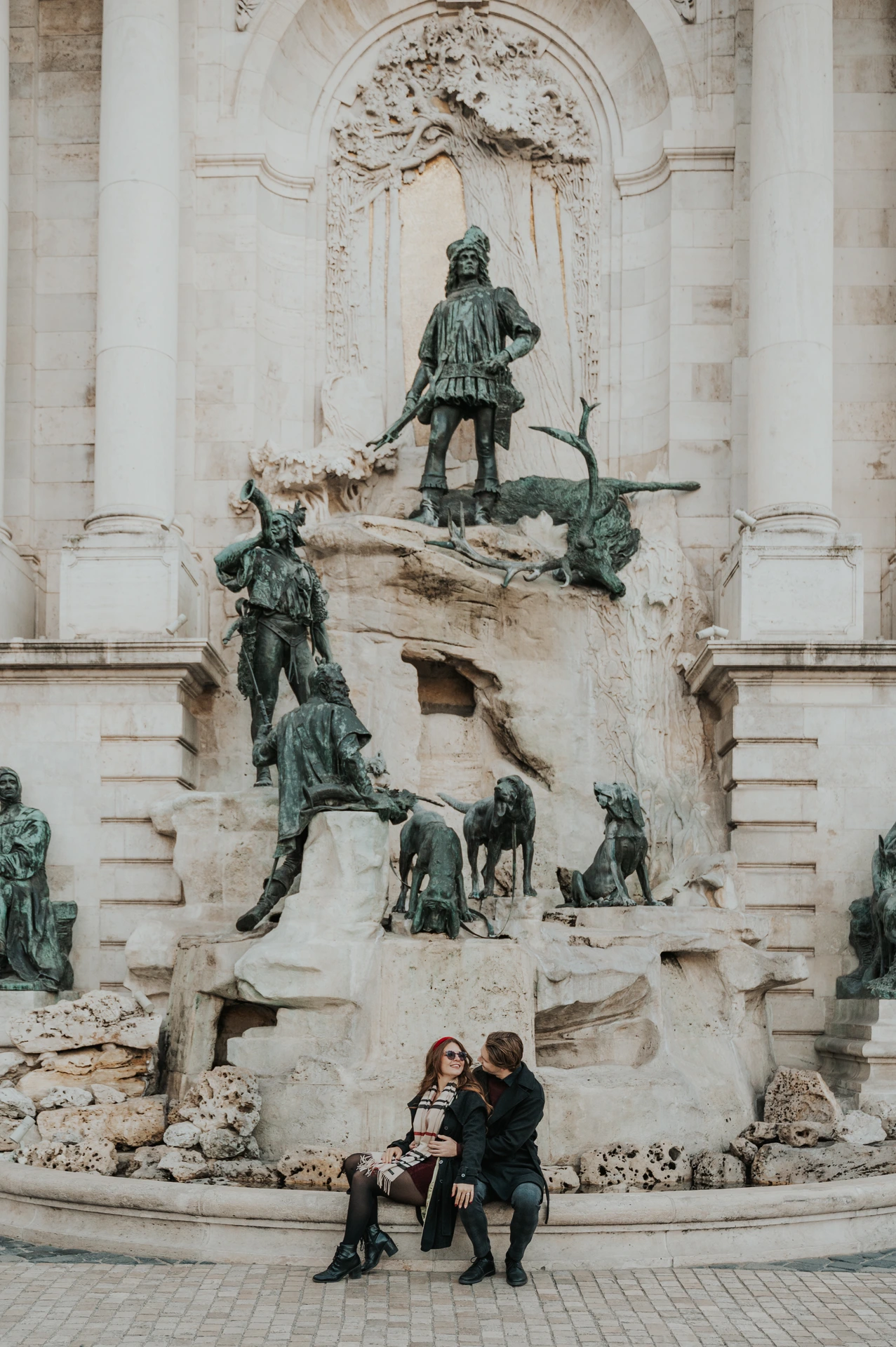 Couple sitting together on a bench beside the sculpted Buda Castle fountain during their Budapest photoshoot
