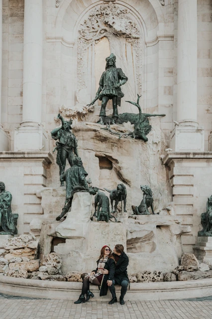 Couple photoshoot in Budapest on a bench by the castle fountain Couple sitting together on a bench beside the sculpted Buda Castle fountain during their Budapest photoshoot