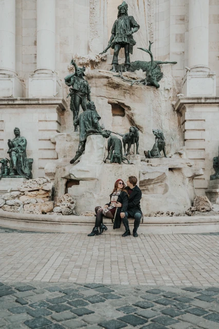 Budapest couple photoshoot with the castle fountain in the backdrop Wide portrait of a couple on a bench beneath the Buda Castle fountain sculptures during their photoshoot in Budapest