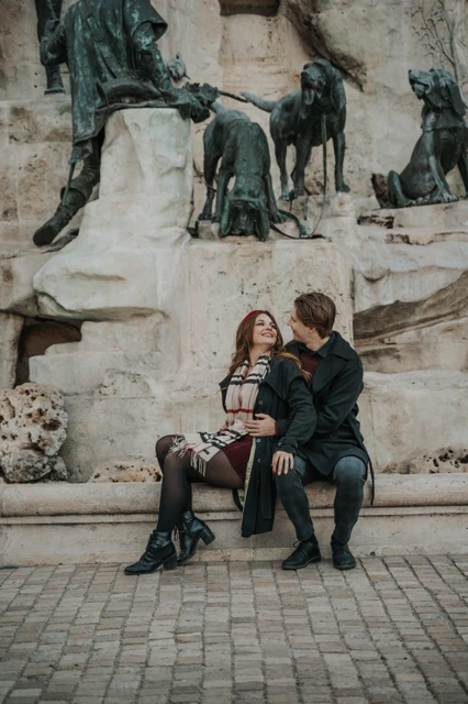 Morning couple photoshoot in Budapest by the castle fountain Couple laughing together on a bench beside the Buda Castle fountain during their morning photoshoot in Budapest