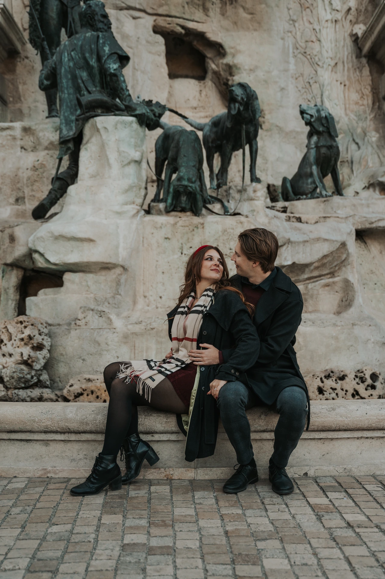Couple looking at each other on a bench beside the fountain sculptures at Buda Castle during their Budapest photoshoot