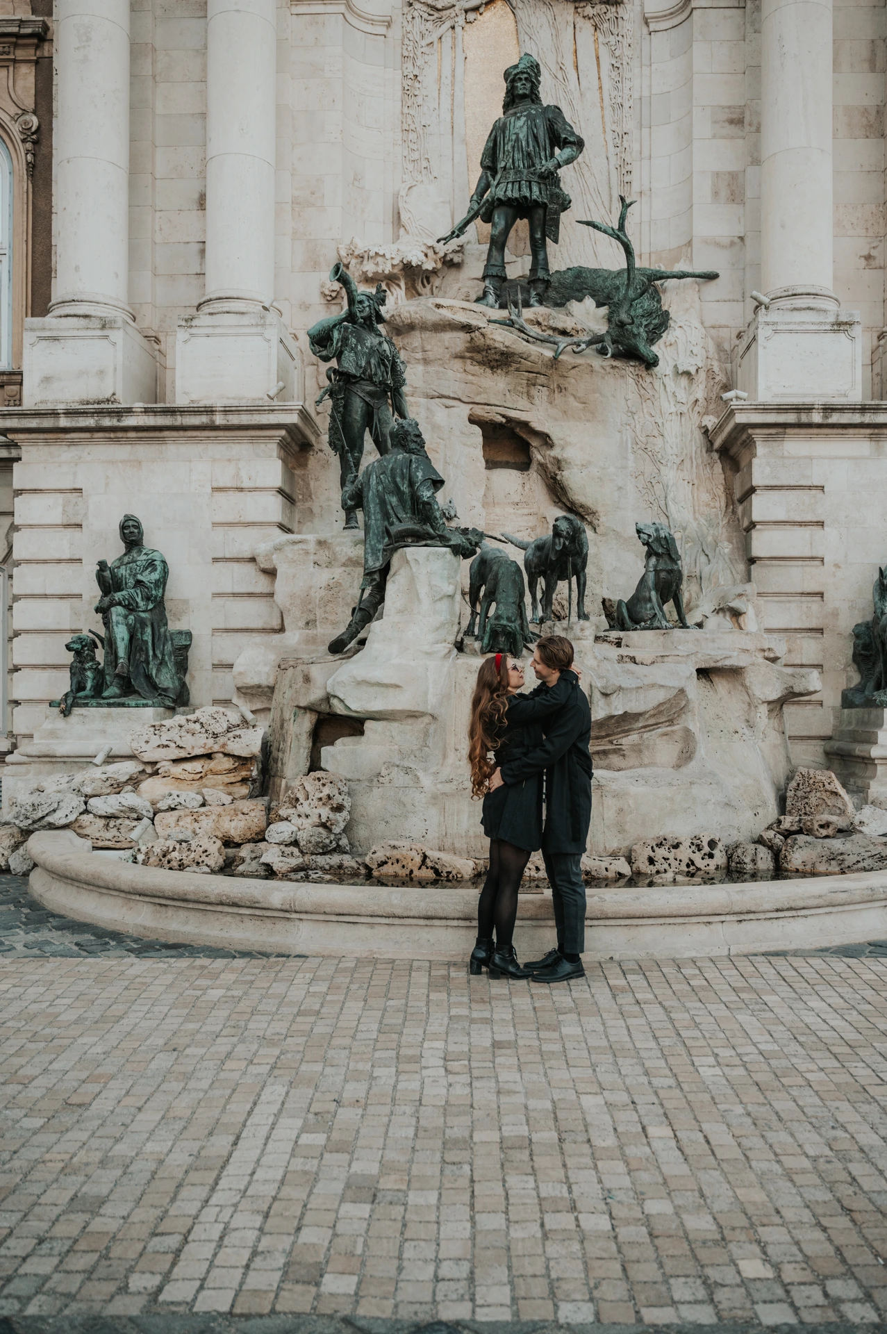 Couple embracing on the cobbled Buda Castle grounds during their Budapest photoshoot, with fountain sculptures behind them