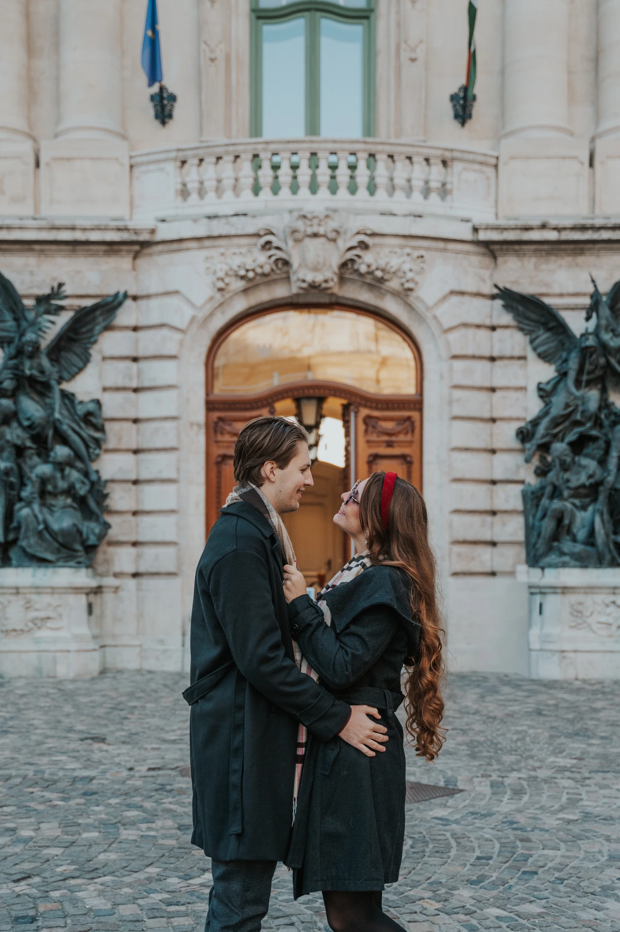 Man kissing his partner on the forehead beside a wooden Buda Castle doorway and bronze statues during their Budapest photoshoot