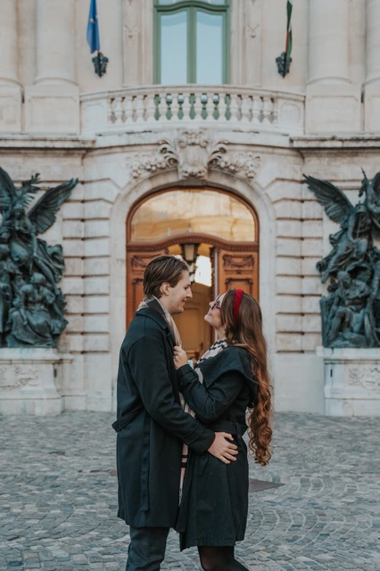 Couple photoshoot in Budapest beside the ornate Buda Castle doorway Man kissing his partner on the forehead beside a wooden Buda Castle doorway and bronze statues during their Budapest photoshoot