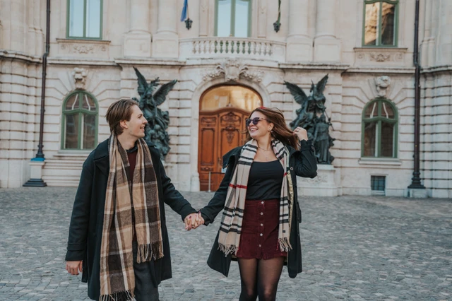 Wide photograph of a couple laughing and holding hands in front of Buda Castle during their couple photoshoot in Budapest