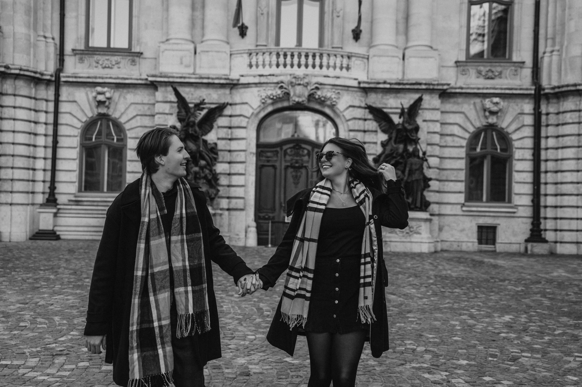 Black and white landscape portrait of a couple laughing and holding hands outside Buda Castle during their Budapest photoshoot