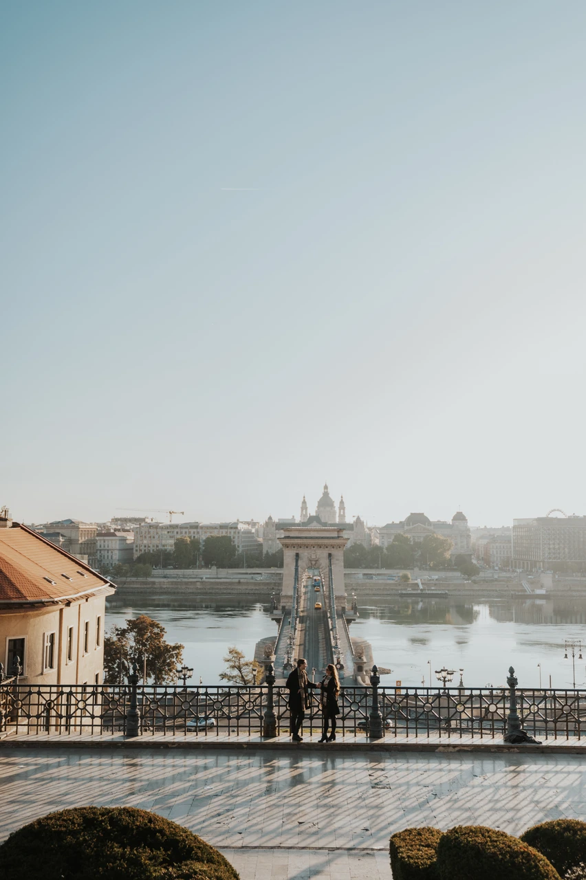 Budapest couple photoshoot overlooking the Chain Bridge Couple standing at the railing of a lower Buda Castle platform during their Budapest photoshoot, with the Chain Bridge and Danube behind them