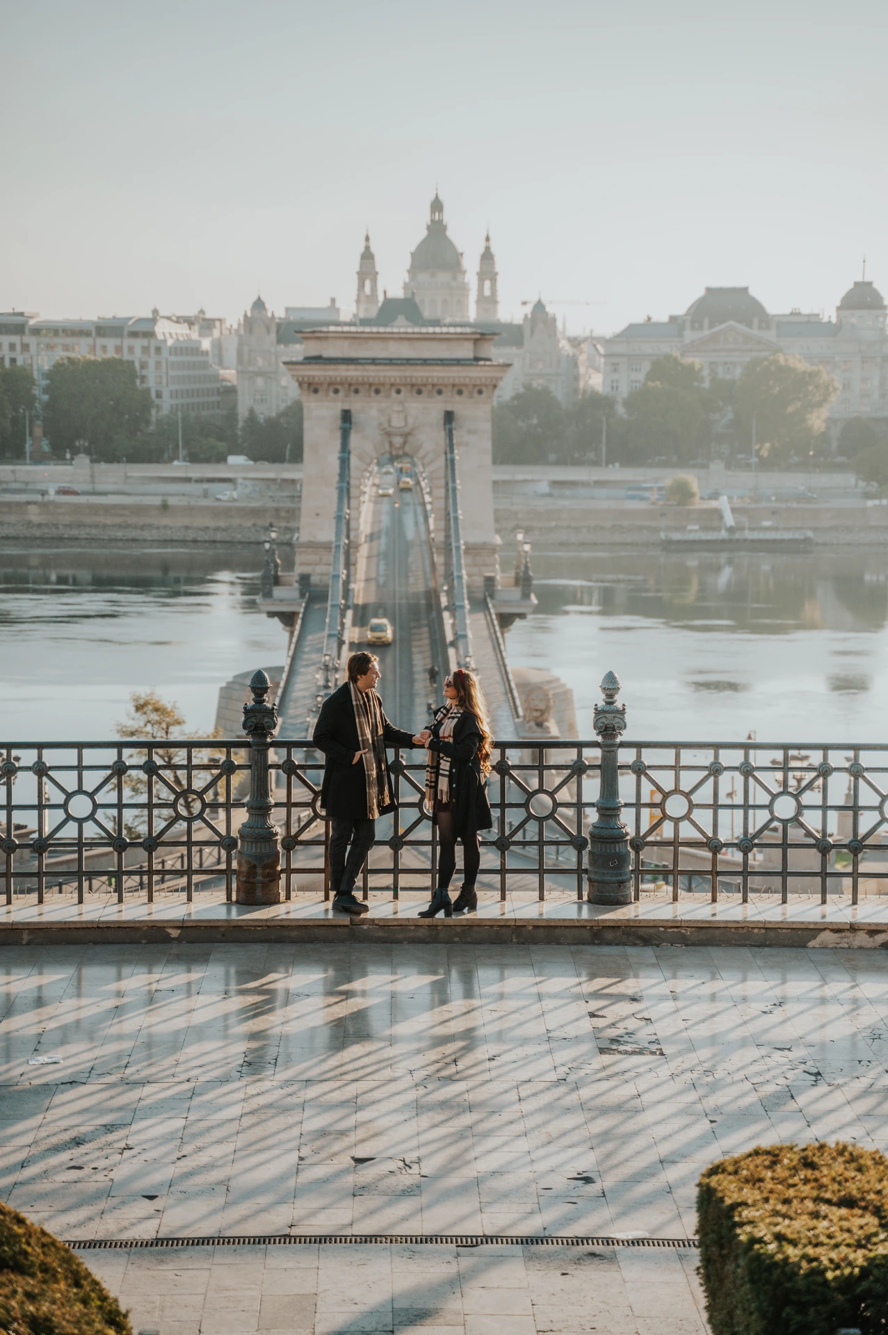 Couple holding hands on the lower Buda Castle platform during their photoshoot in Budapest, with the Chain Bridge in the background