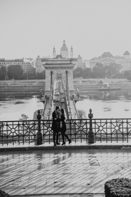 Black and white photoshoot in Budapest with the Chain Bridge in the backdrop Black and white portrait of a couple at a lower Buda Castle viewpoint during their Budapest photoshoot, with the Chain Bridge behind them