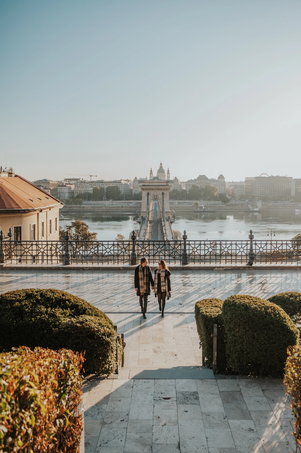 Couple walking along a lower Buda Castle platform during their Budapest photoshoot, with the Chain Bridge and Danube in the background