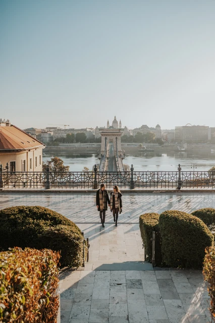 Walking couple photoshoot in Budapest with the Chain Bridge in the background Couple walking along a lower Buda Castle platform during their Budapest photoshoot, with the Chain Bridge and Danube in the background
