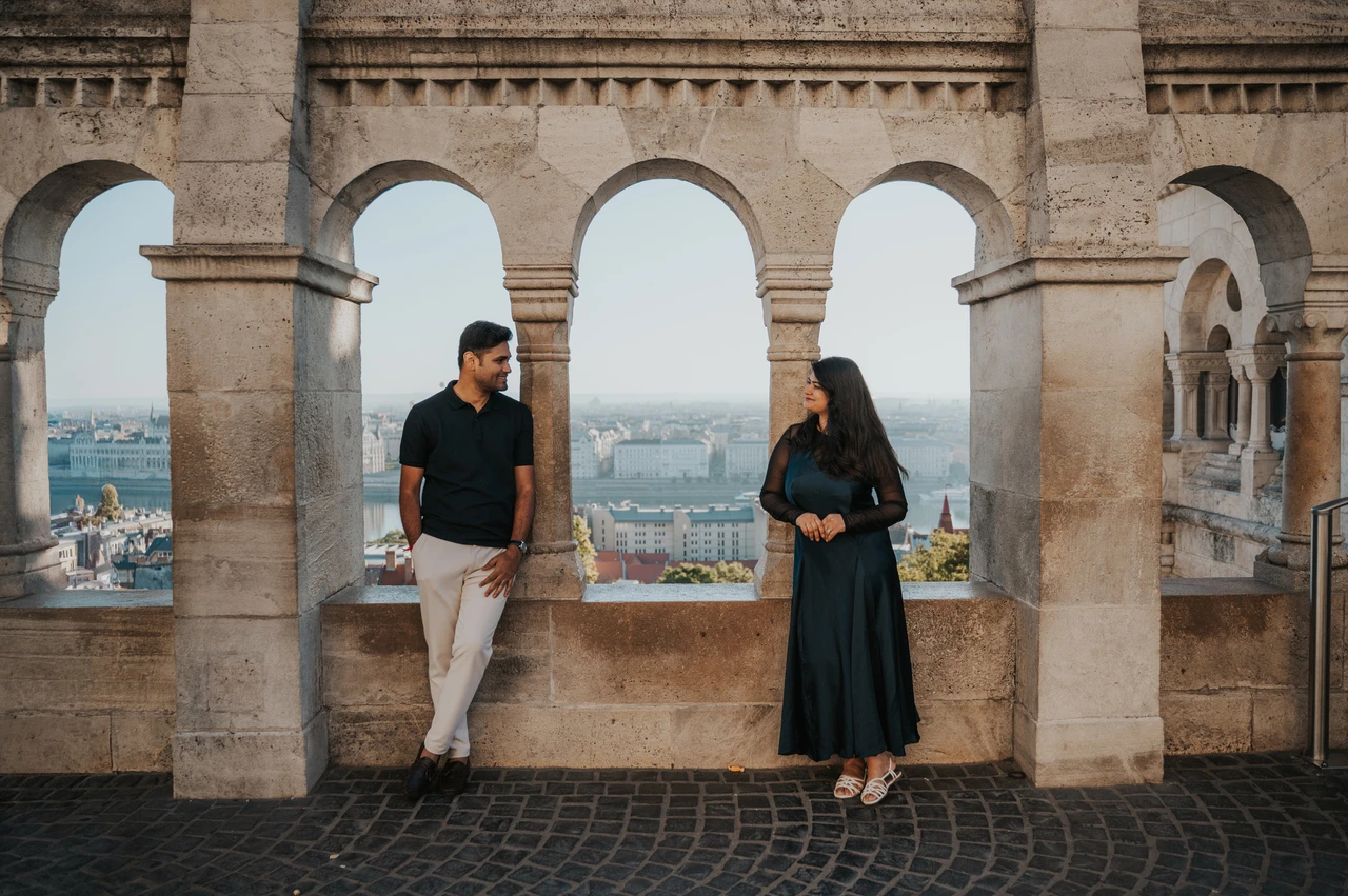 Budapest couple photoshoot: a wide view through the Bastion’s arches A wide portrait of the couple standing apart between the stone arches at Fisherman’s Bastion during a couple photoshoot in Budapest.