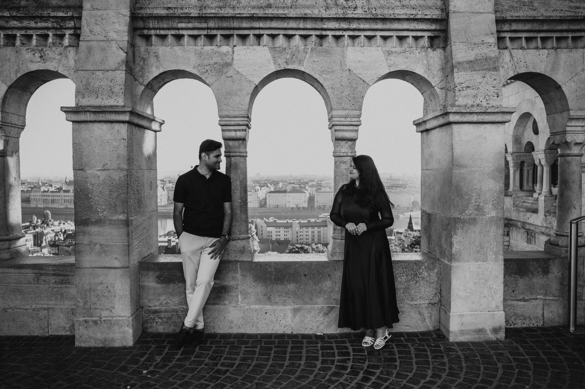 Black-and-white photograph of the couple standing at separate stone arches with the city beyond Fisherman’s Bastion during a couple photoshoot in Budapest.