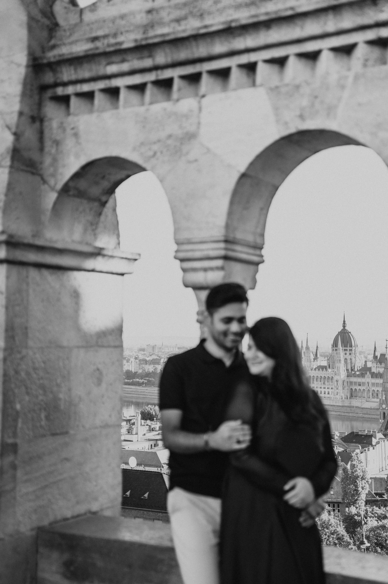 Black-and-white photograph of the couple leaning close together beside the open arcade at Fisherman’s Bastion during a couple photoshoot in Budapest.