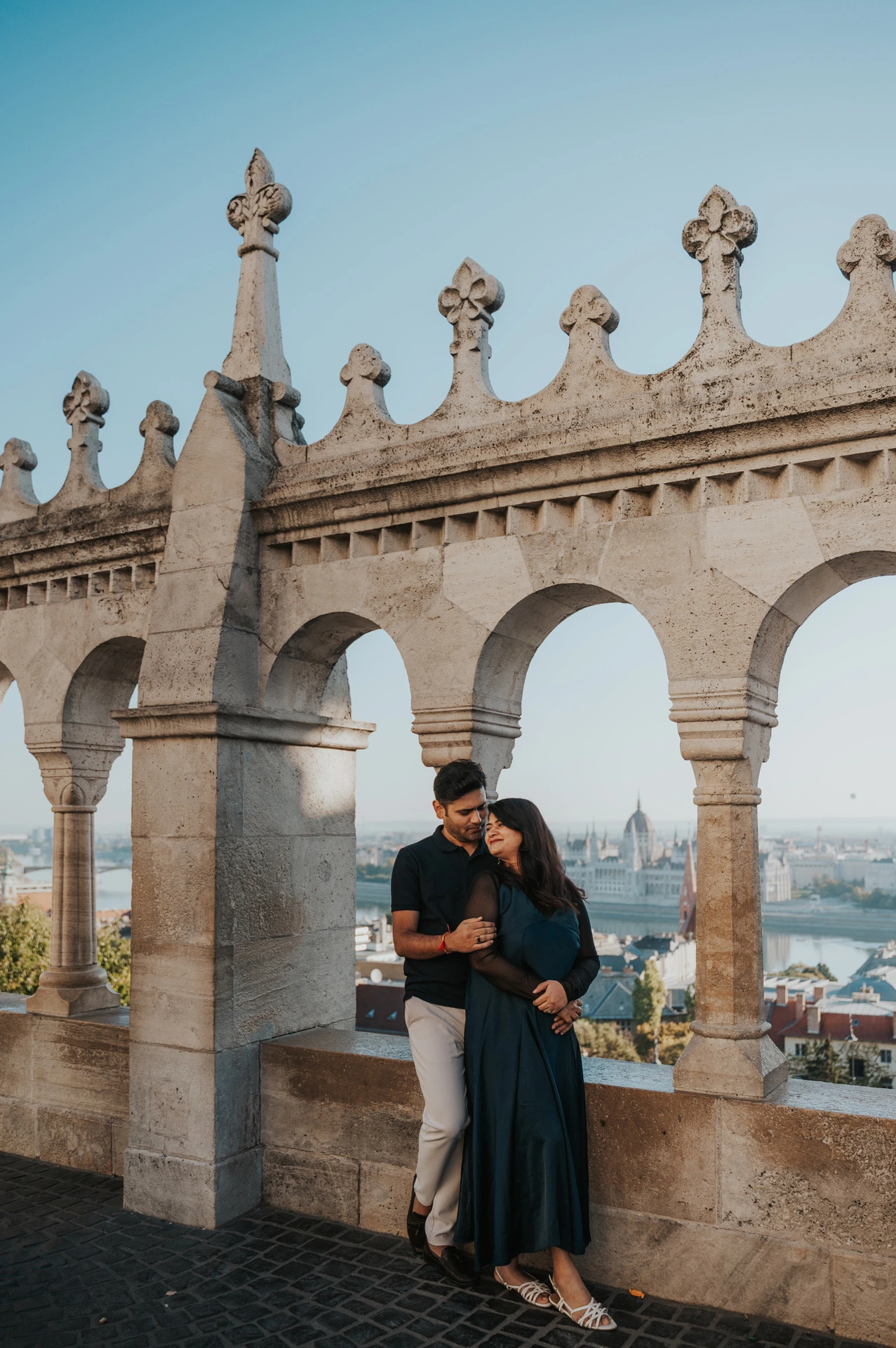 The couple leaning close beside the open stone arches and decorative parapet at Fisherman’s Bastion during a couple photoshoot in Budapest.