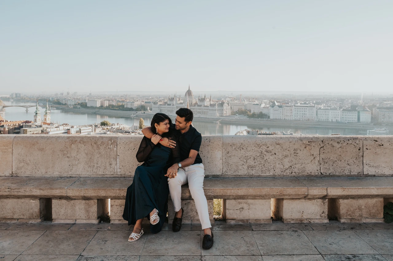 Budapest couple photoshoot: a wide portrait on the Bastion’s stone bench A landscape portrait of the couple seated on a stone bench with the city behind them during a couple photoshoot in Budapest.