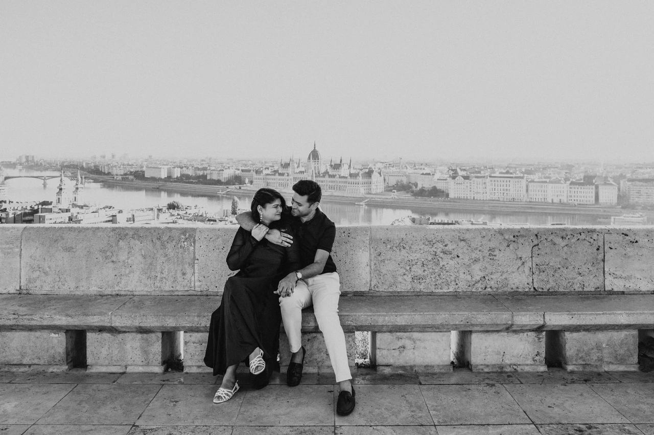 Budapest couple photoshoot: a bench portrait with the city beyond in black and white Black-and-white photograph of the couple seated close on a stone bench overlooking the city from Fisherman’s Bastion during a couple photoshoot in Budapest.