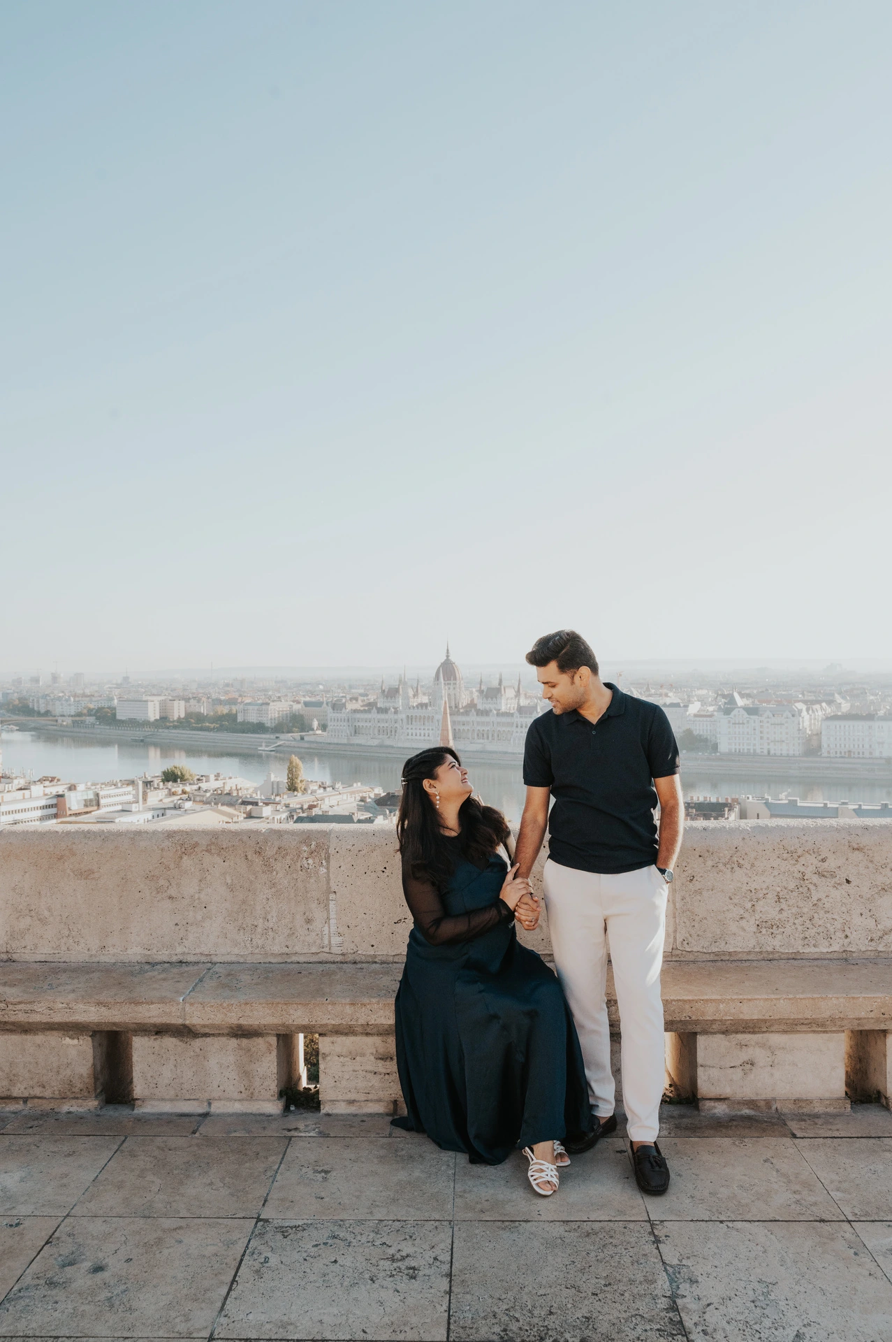 The woman sitting on a stone bench while her partner stands beside her at Fisherman’s Bastion during a couple photoshoot in Budapest.