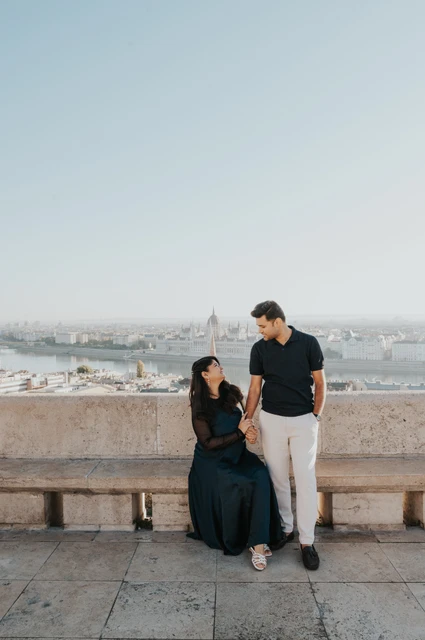 Budapest couple photoshoot: a seated portrait beside her partner The woman sitting on a stone bench while her partner stands beside her at Fisherman’s Bastion during a couple photoshoot in Budapest.
