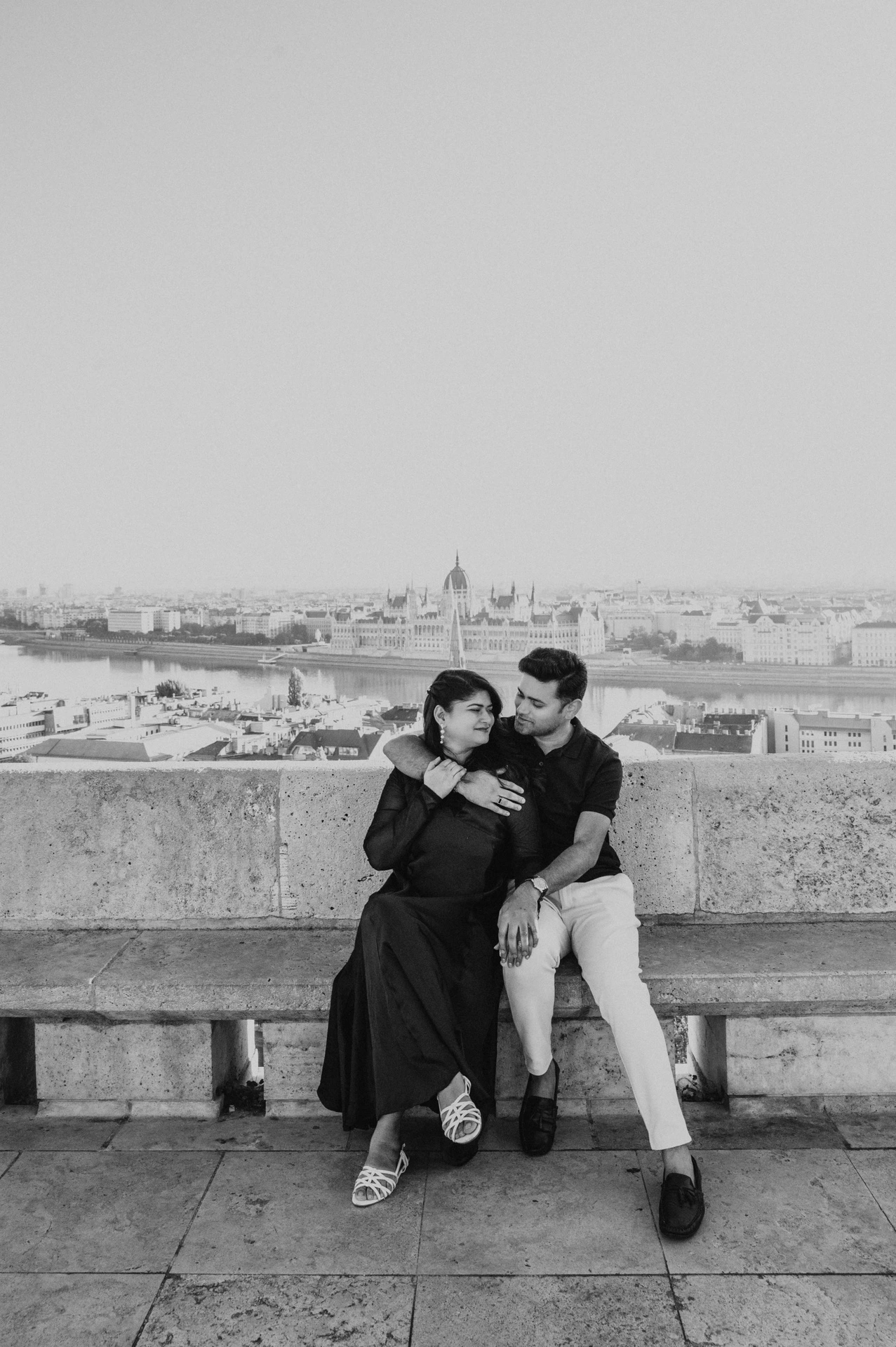 Black-and-white photograph of the couple sitting together on a stone bench overlooking the city from Fisherman’s Bastion during a couple photoshoot in Budapest.