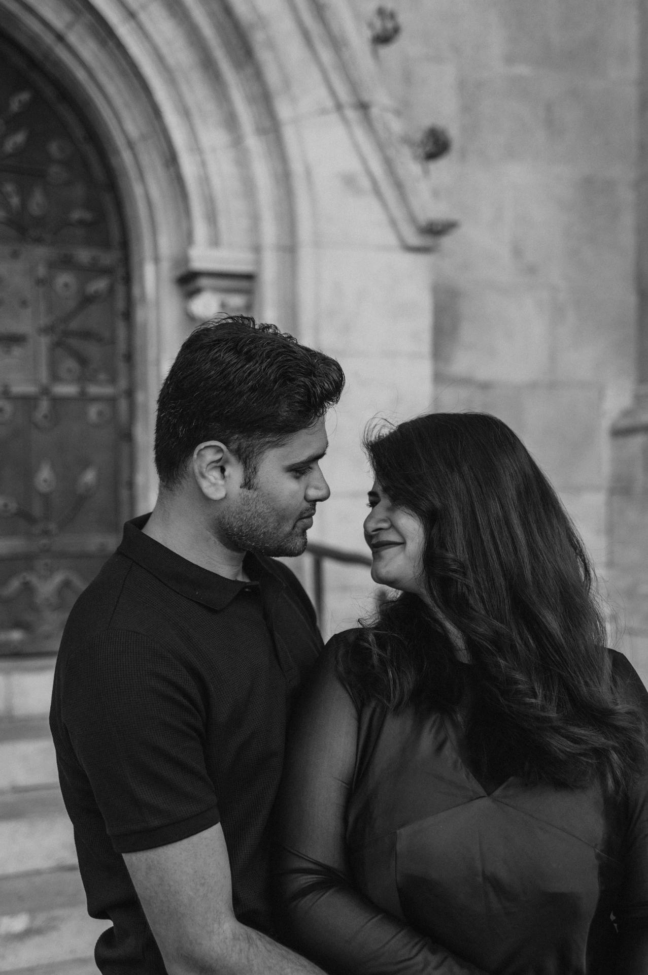 Black-and-white photograph of the couple smiling towards each other in front of the pointed arch at Matthias Church during a couple photoshoot in Budapest.