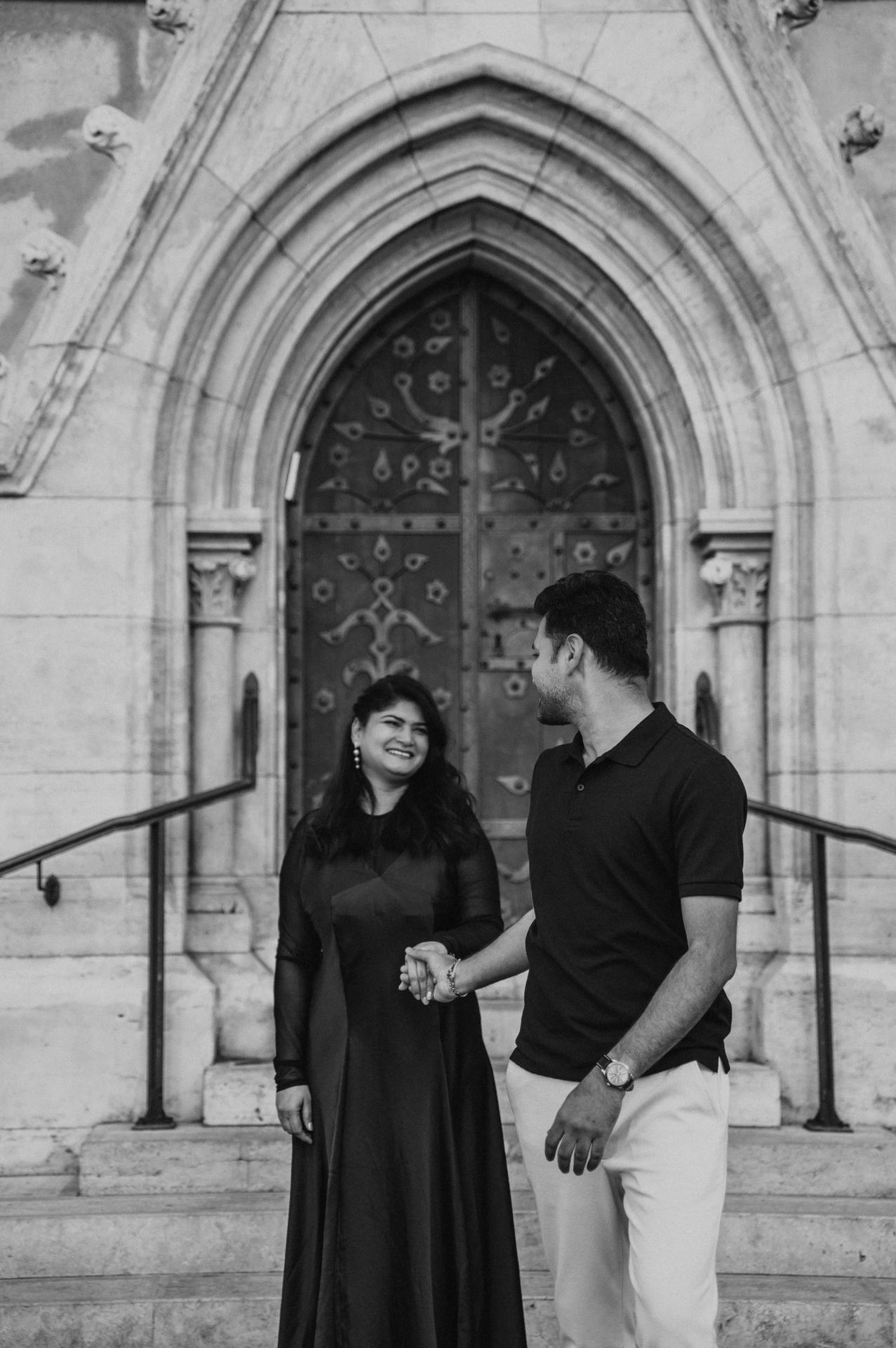 Black-and-white photograph of the couple looking towards each other beside a carved church doorway and stone steps during a couple photoshoot in Budapest.