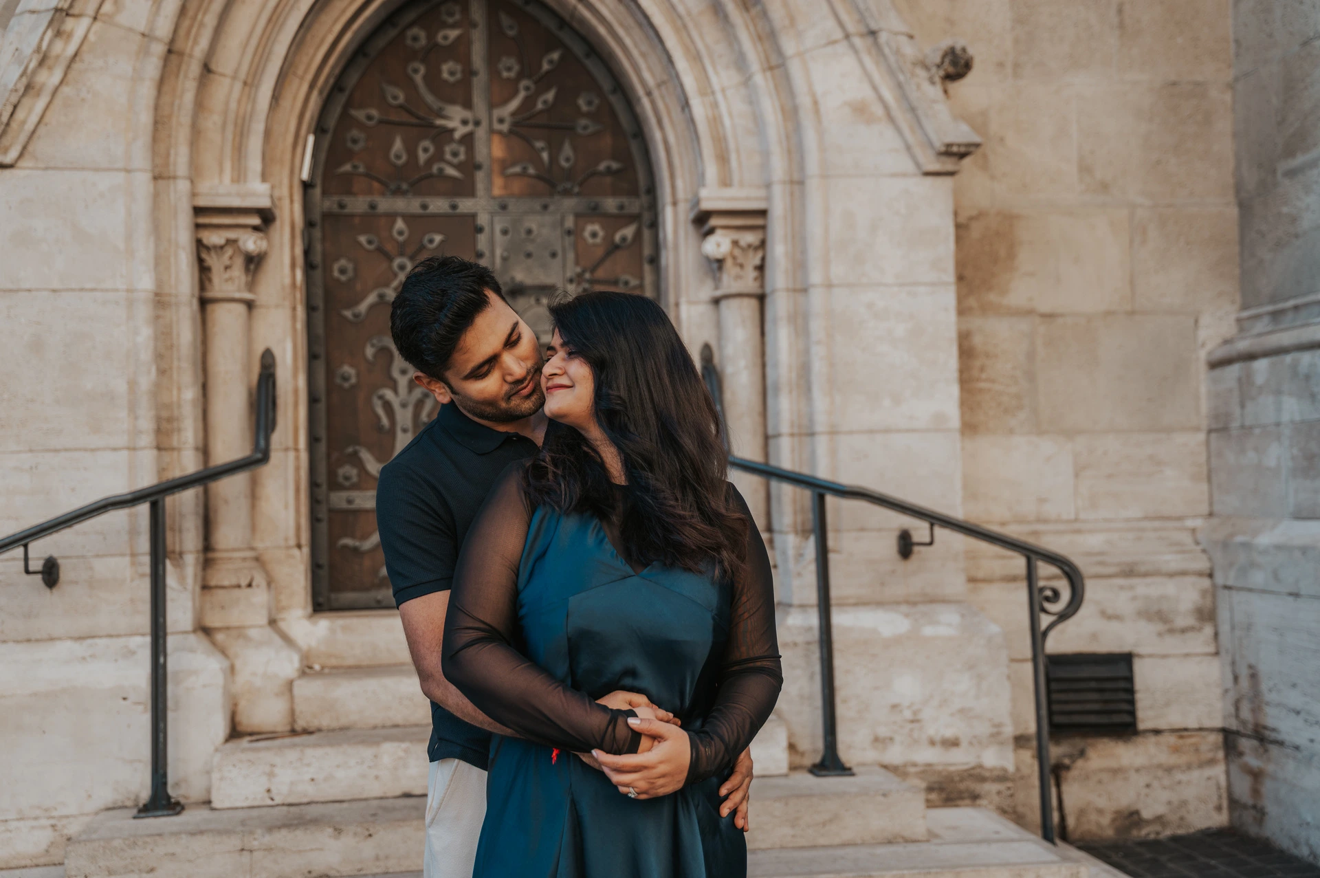 A landscape portrait of the man kissing his partner near her temple by the Matthias Church doorway during a couple photoshoot in Budapest.