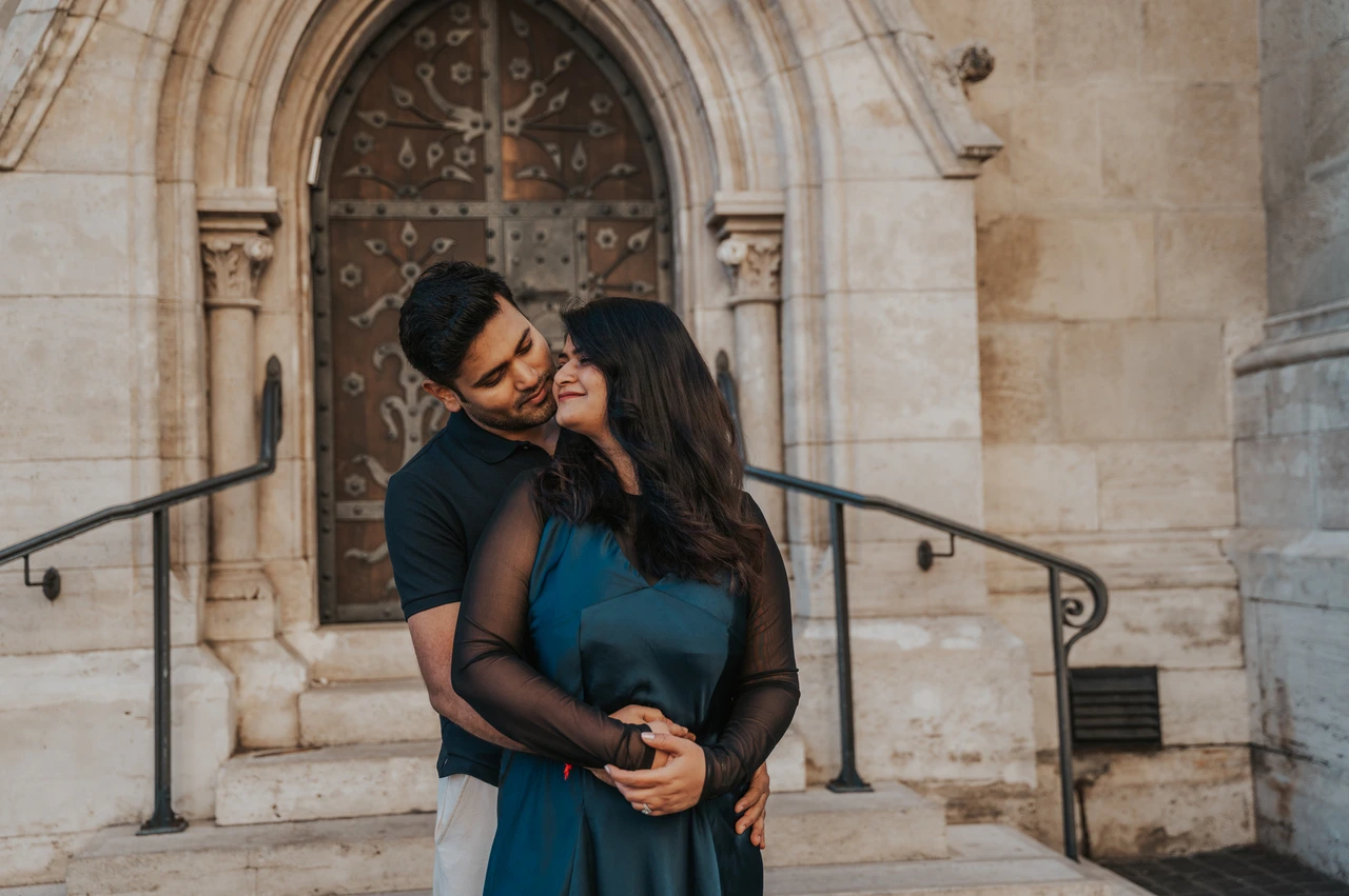 Budapest couple photoshoot: a kiss beside the carved church door A landscape portrait of the man kissing his partner near her temple by the Matthias Church doorway during a couple photoshoot in Budapest.