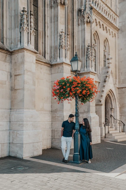 Budapest couple photoshoot: red flowers beside Matthias Church The couple standing beside Matthias Church’s carved stone wall and a lamp with red flowers during a couple photoshoot in Budapest.