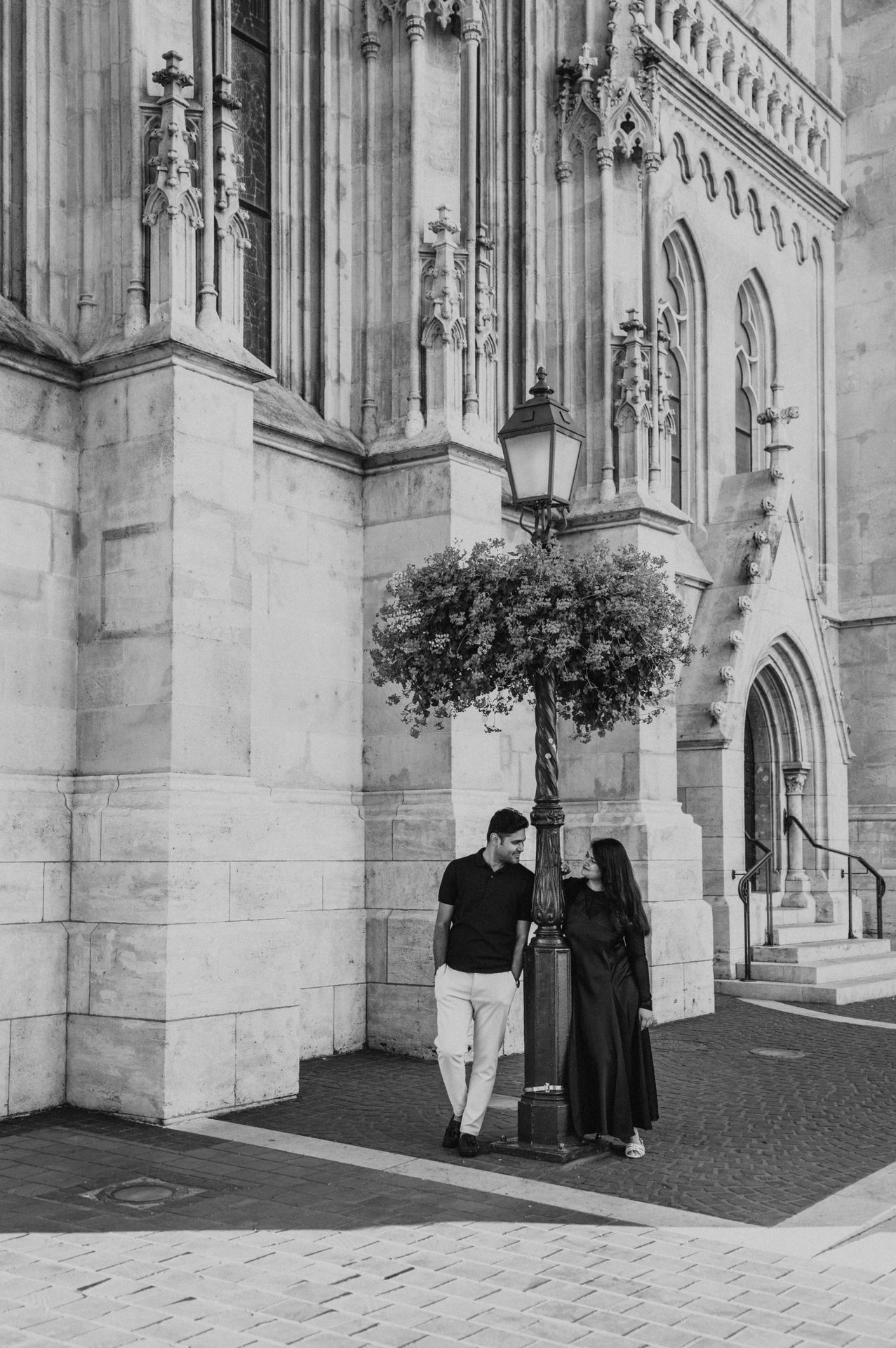 Black-and-white photograph of the couple beside a flower-hung lamp and carved stonework at Matthias Church during a couple photoshoot in Budapest.